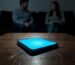 A glowing smartphone on a table with a distant couple in the background, symbolizing digital cheating and the need for online affair recovery.